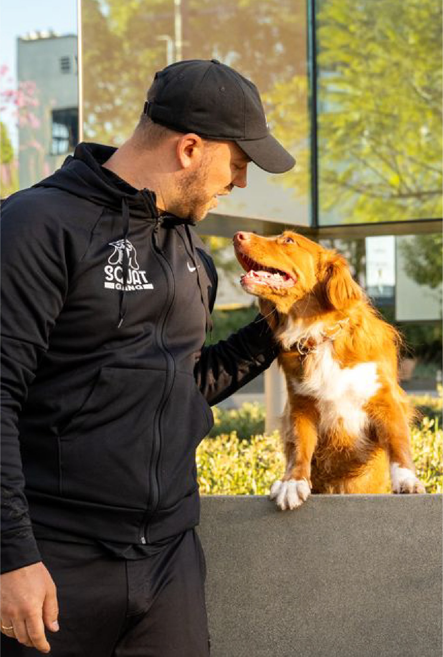 A man in a black jacket interacts affectionately with a brown dog outdoors.