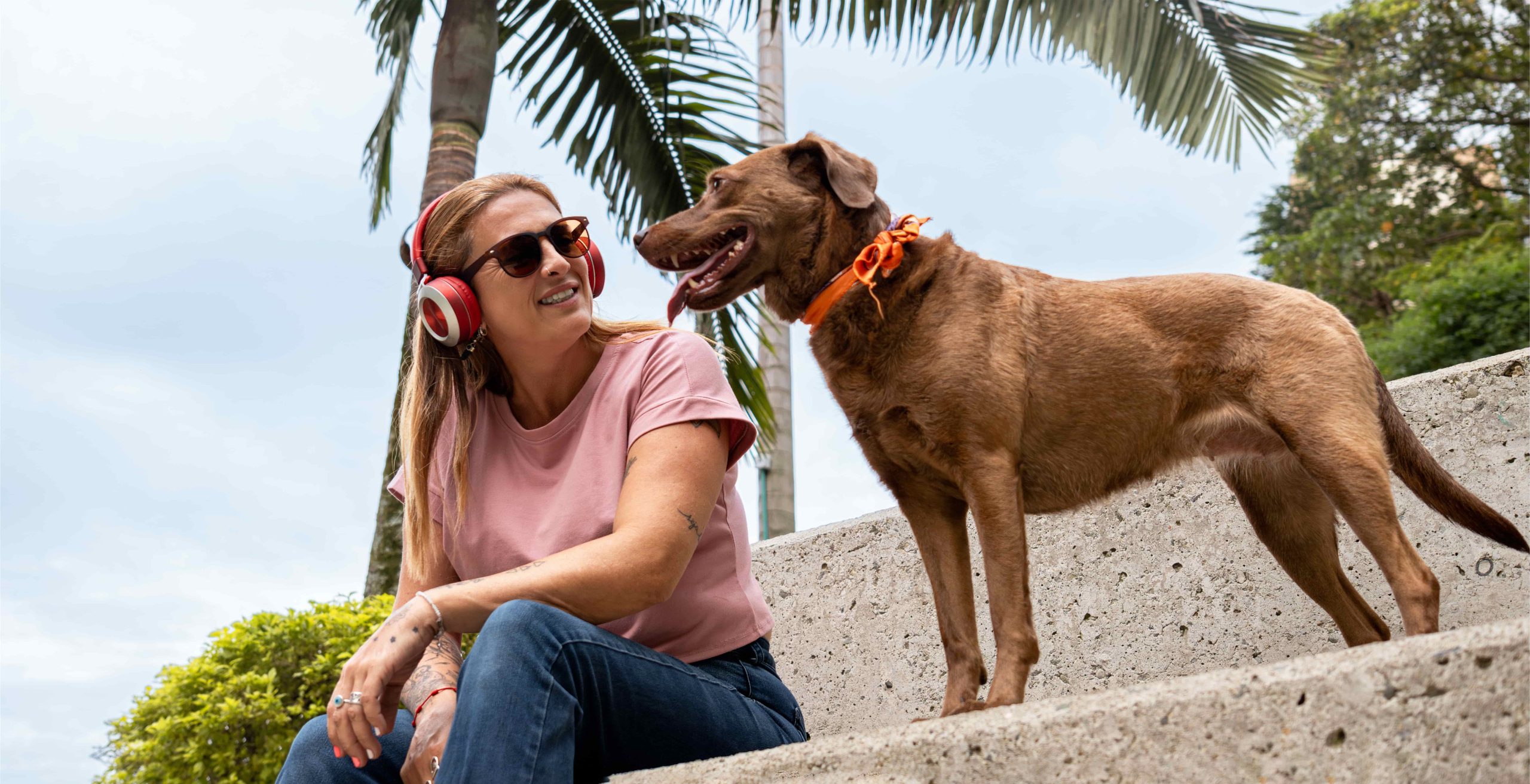 Woman with headphones smiles at a brown dog wearing an orange collar on a concrete step.