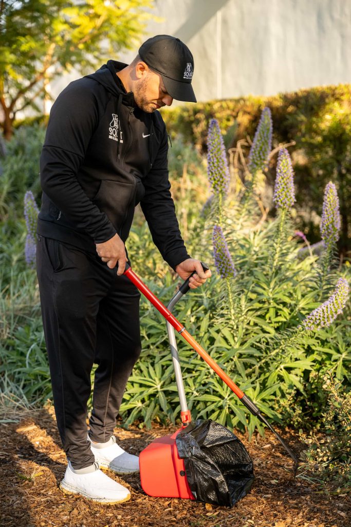 Man in a black outfit using a gardening tool near colorful plants and mulch.